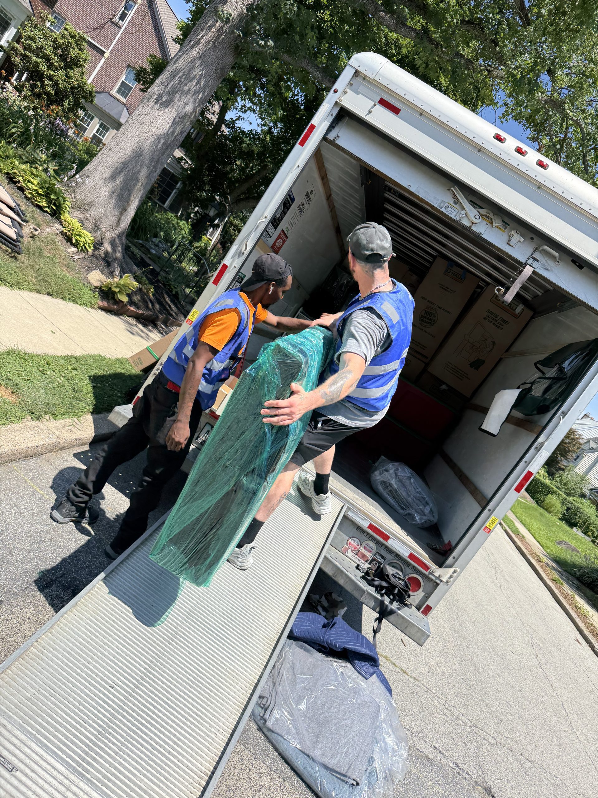 Two professional movers from A+ Cleaning & Moving loading shrink-wrapped furniture into a moving truck in a suburban Delaware County neighborhood.